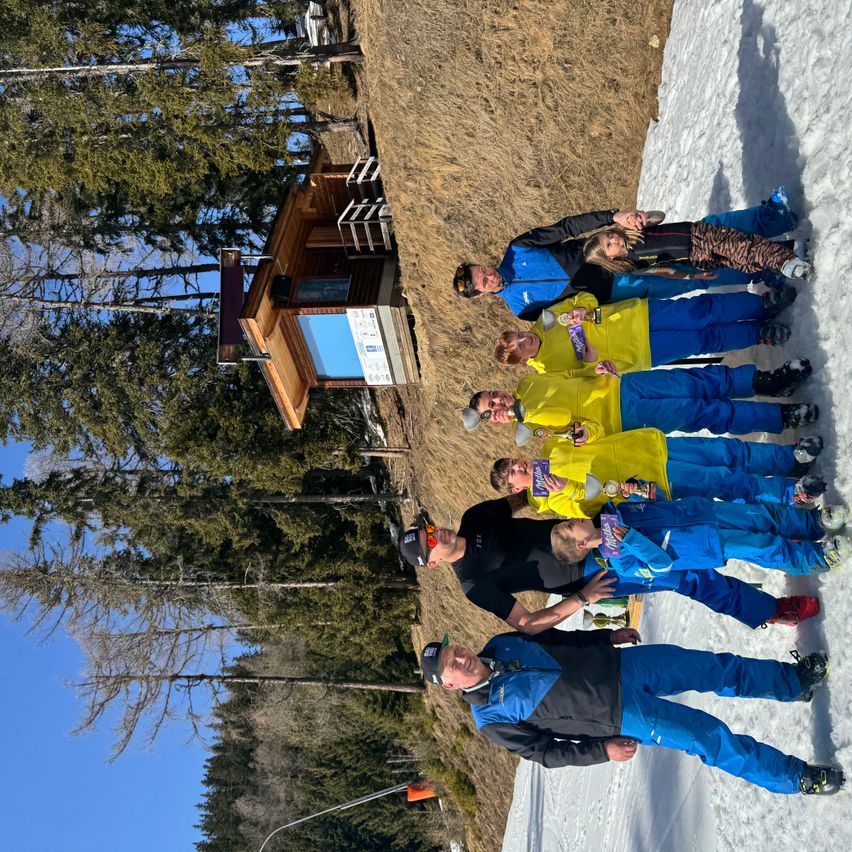 Eine Gruppe von Skifahrern posiert für ein Foto auf einer verschneiten Piste, mit einer kleinen Holzstruktur im Hintergrund.