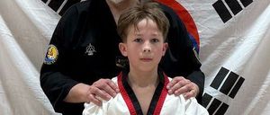 Ein Mann und ein junger Junge stehen in einem Kampfsportstudio, beide tragen schwarze und weiße Uniformen. Der Mann hat seine Hände auf den Schultern des Jungen. Hinter ihnen befindet sich ein weißer Banner mit schwarzen Symbolen.