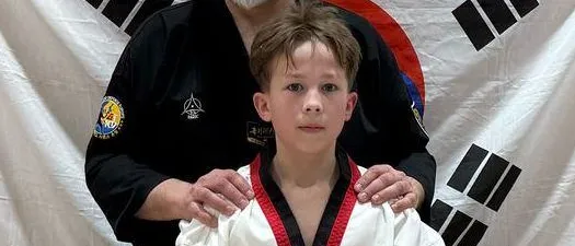 Ein Mann und ein junger Junge stehen in einem Kampfsportstudio, beide tragen schwarze und weiße Uniformen. Der Mann hat seine Hände auf den Schultern des Jungen. Hinter ihnen befindet sich ein weißer Banner mit schwarzen Symbolen.