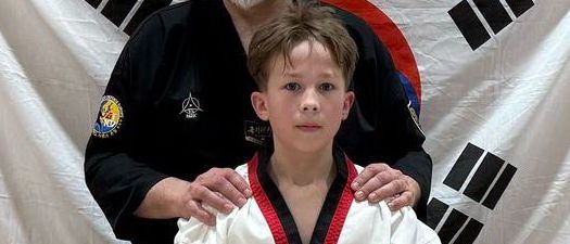 Ein Mann und ein junger Junge stehen in einem Kampfsportstudio, beide tragen schwarze und weiße Uniformen. Der Mann hat seine Hände auf den Schultern des Jungen. Hinter ihnen befindet sich ein weißer Banner mit schwarzen Symbolen.