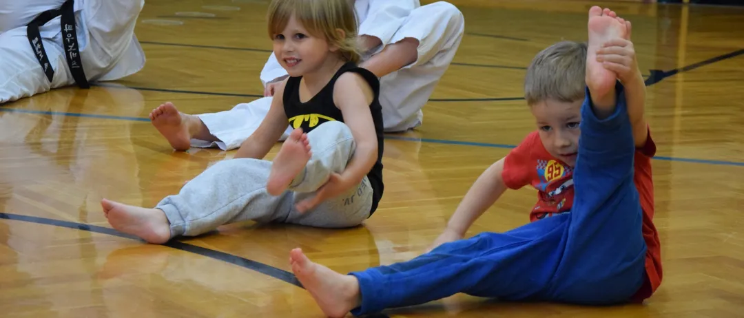 Drei kleine Kinder sitzen auf einem Holzboden, der wie eine Turnhalle aussieht. Ein Kind mit einem Batman-Shirt lächelt und streckt die Beine.