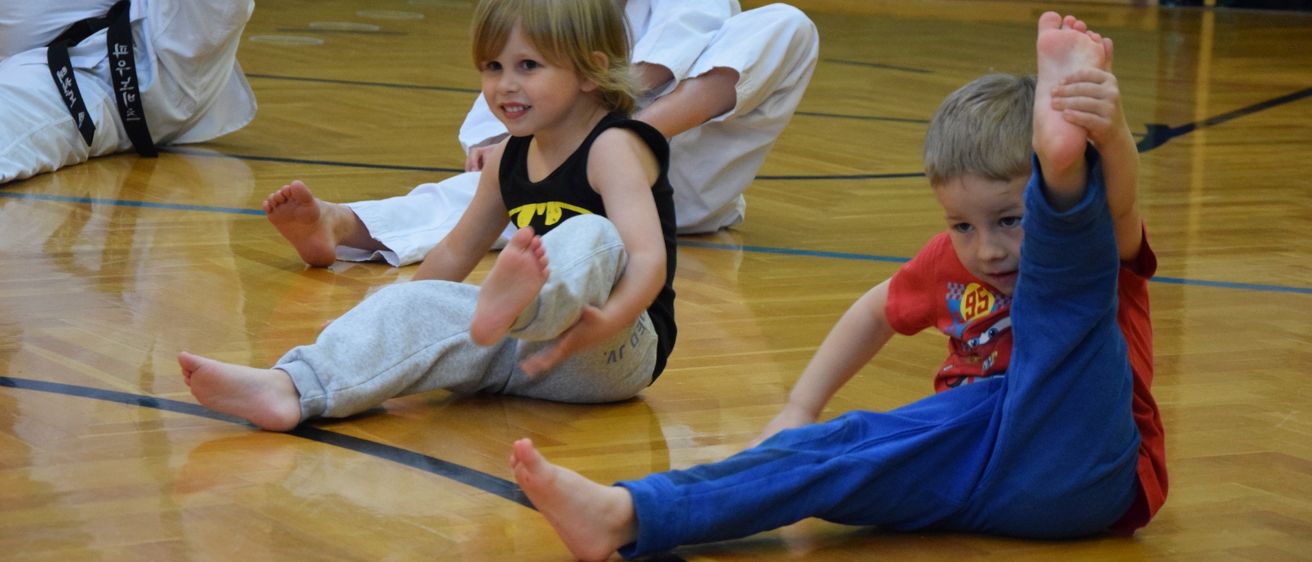 Drei kleine Kinder sitzen auf einem Holzboden, der wie eine Turnhalle aussieht. Ein Kind mit einem Batman-Shirt lächelt und streckt die Beine.