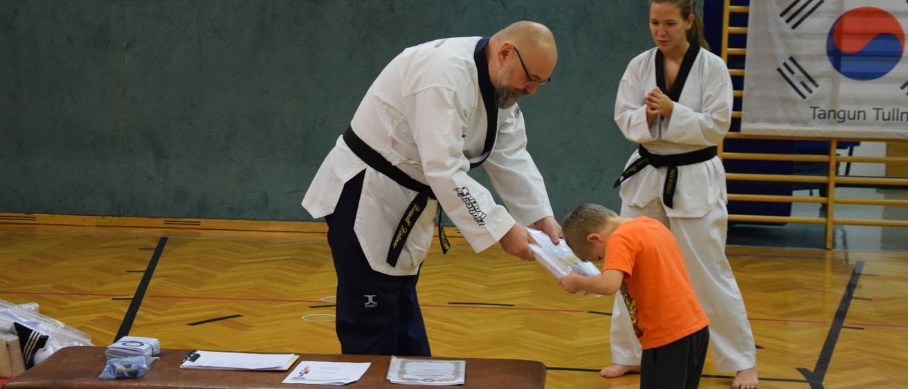 In einer Turnhalle überreicht ein Karatelehrer einem jungen Jungen, der auf das Papier schaut, ein Zertifikat. Ein Mädchen in einer Karateuniform steht hinter ihnen.