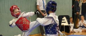 Zwei junge Kinder in Schutzkleidung üben Kampfsport auf einem blauen Teppich. Ein Mann in Krawatte beobachtet sie.