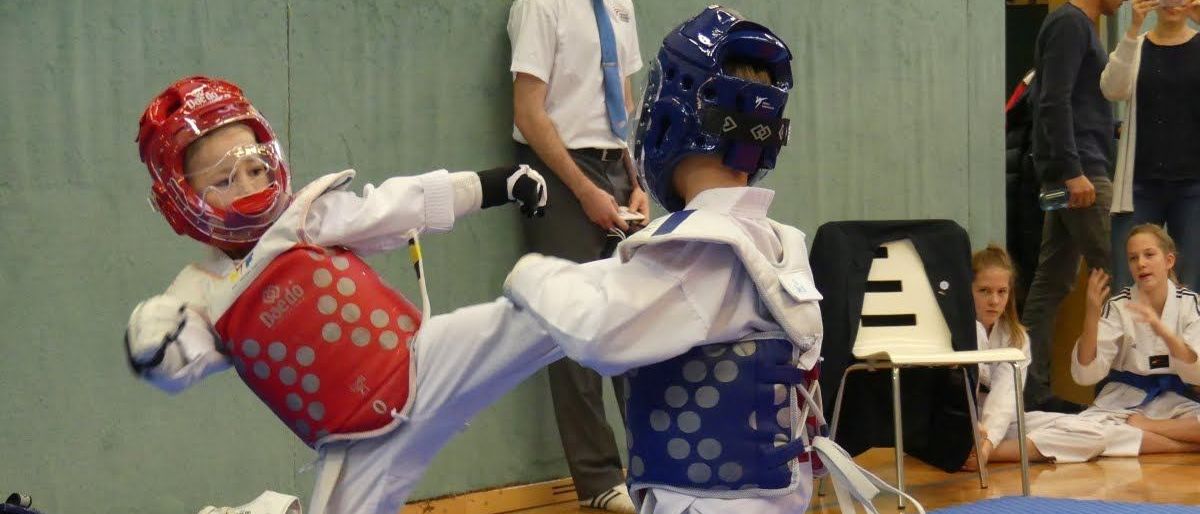 Zwei junge Kinder in Schutzkleidung üben Kampfsport auf einem blauen Teppich. Ein Mann in Krawatte beobachtet sie.