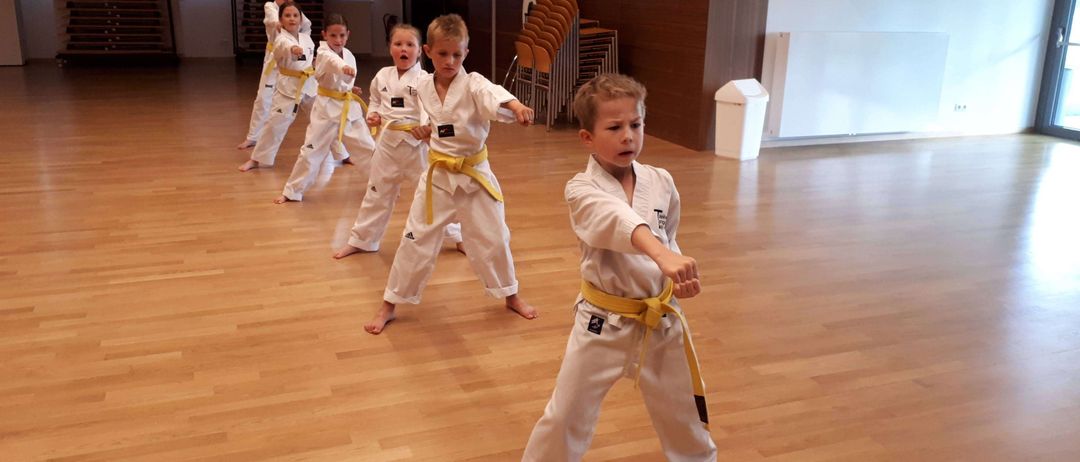 Eine Gruppe junger Kinder in weißen Karate-Uniformen mit gelben Gürteln übt Karate in einem Raum.