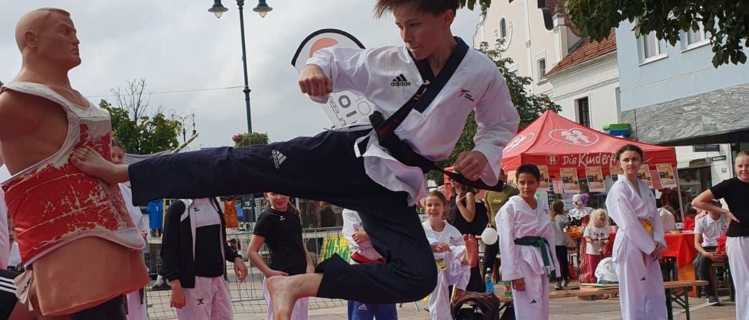 Ein Kampfsportschüler tritt einen Dummy auf einem öffentlichen Platz, mit Zuschauern, einem Zelt und einer Straßenlaterne im Hintergrund.