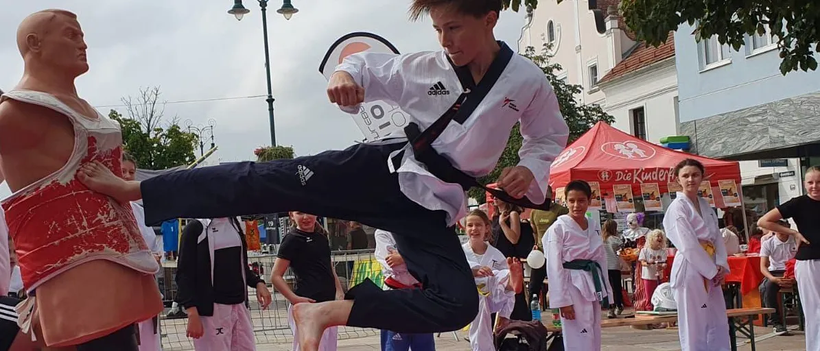 Ein Kampfsportschüler tritt einen Dummy auf einem öffentlichen Platz, mit Zuschauern, einem Zelt und einer Straßenlaterne im Hintergrund.