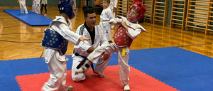 In einem Kampfsportkurs übt ein junger Junge mit rotem Helm einen Tritt mit einem Mann im schwarzen Gürtel, während ein anderer Junge mit blauem Helm zusieht.