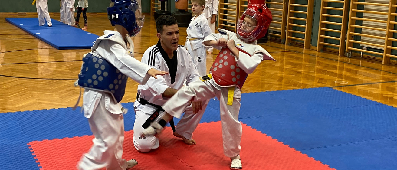 In einem Kampfsportkurs übt ein junger Junge mit rotem Helm einen Tritt mit einem Mann im schwarzen Gürtel, während ein anderer Junge mit blauem Helm zusieht.