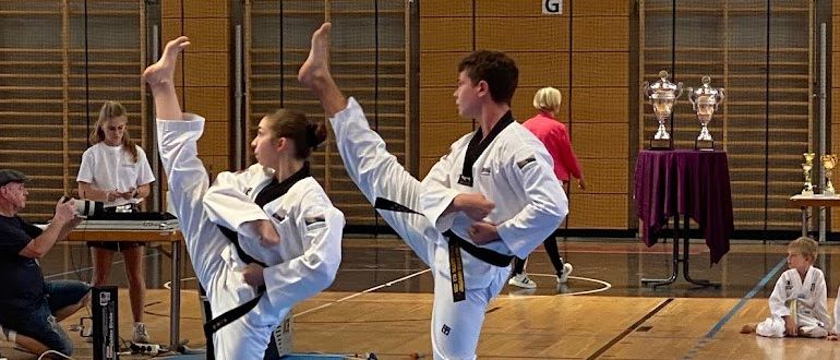 Zwei Personen in weißen Uniformen führen Kampfkunst-Tritte in einer Turnhalle aus. Dahinter ist ein Tisch mit Trophäen zu sehen. Zuschauer und Teilnehmer sind anwesend, einige sitzend, andere stehend. Die Turnhalle hat einen blauen Boden und Glaswände.