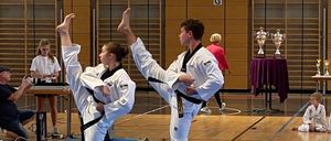 Zwei Personen in weißen Uniformen führen Kampfkunst-Tritte in einer Turnhalle aus. Dahinter ist ein Tisch mit Trophäen zu sehen. Zuschauer und Teilnehmer sind anwesend, einige sitzend, andere stehend. Die Turnhalle hat einen blauen Boden und Glaswände.