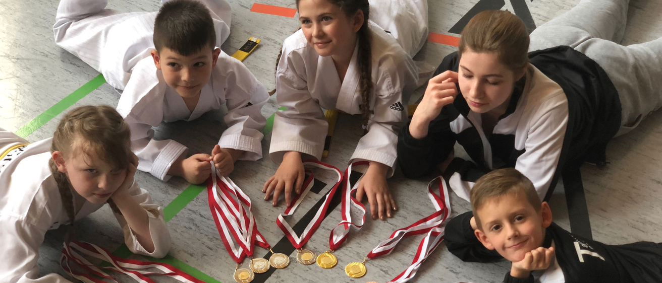 Fünf Kinder in Karate-Uniformen liegen lächelnd auf dem Boden, mit Medaillen um den Hals und in den Händen. Die Medaillen sind gold und silber, mit Bändern. Der Boden hat grüne und rote Linien.