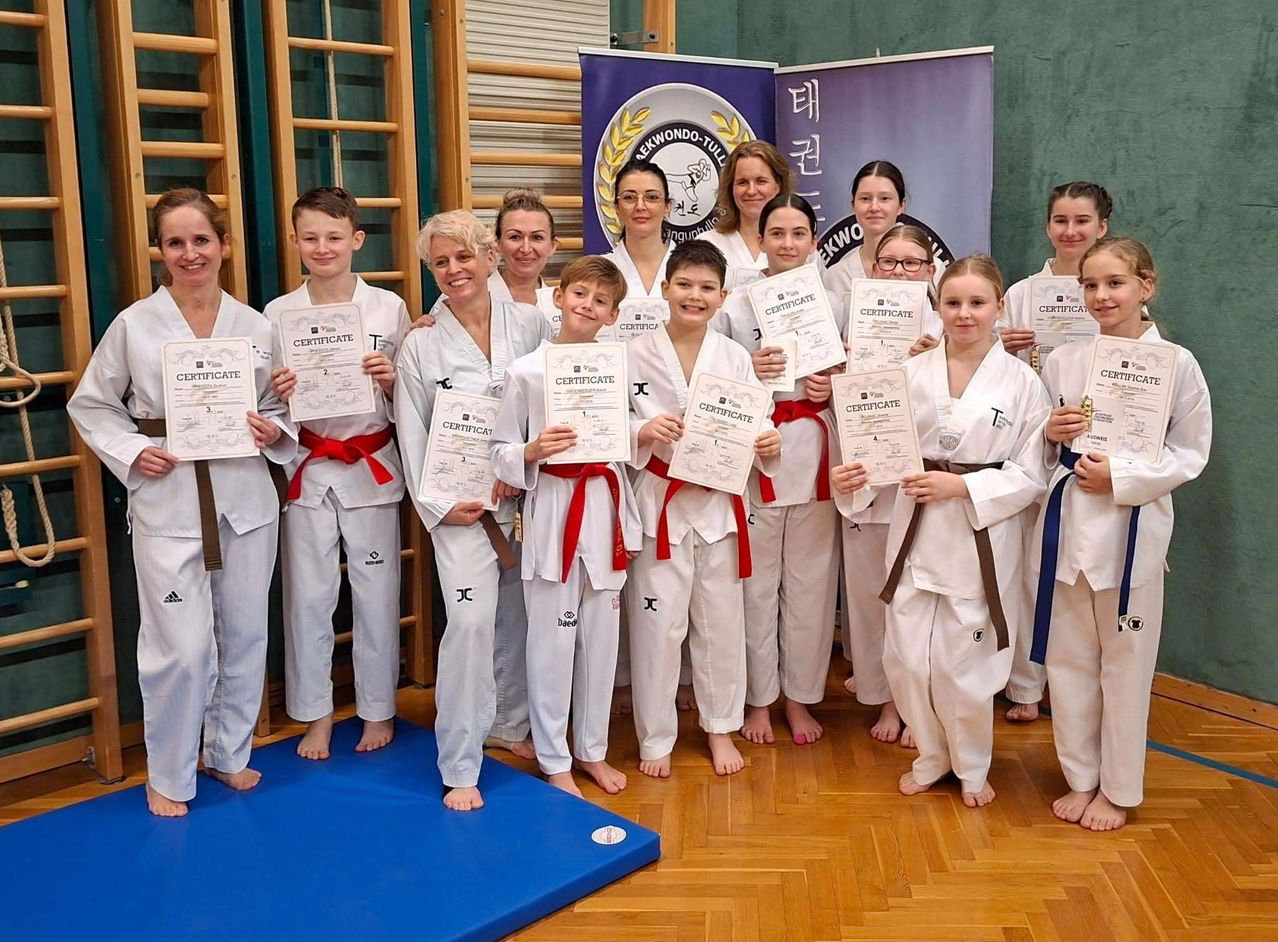 Eine Gruppe junger Kampfsportler, darunter Jungen und Mädchen, halten Urkunden in den Händen und posieren für ein Foto. Sie tragen weiße Uniformen mit roten und blauen Gürteln. Die Urkunden, die sie halten, haben den Text 'Zertifikat' darauf. Hinter ihnen befinden sich zwei Banner mit Logos und Text.