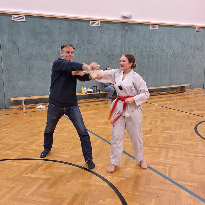 In einer Turnhalle üben ein Mann und eine Frau Kampfsport. Die Frau hält ein Holzschwert, während der Mann ihre Arme hält. Sie sind barfuß auf dem Holzboden.