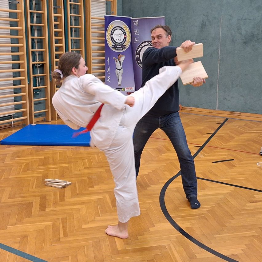 Eine Frau in einem weißen Karate-Anzug tritt einen Mann, der Holzbretter hält, in einer Turnhalle.