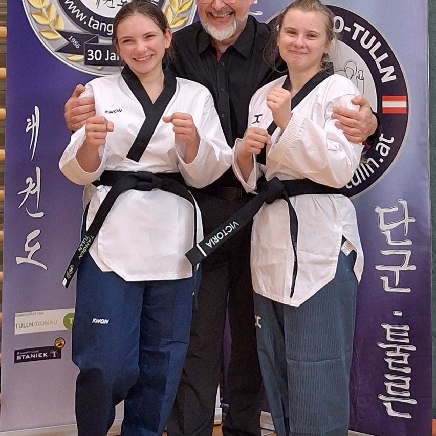 Zwei Frauen in weißen Karate-Uniformen posieren mit einem lächelnden Mann, wahrscheinlich einem Trainer, vor einem Banner mit einem Logo. Die Frauen zeigen Daumen hoch.