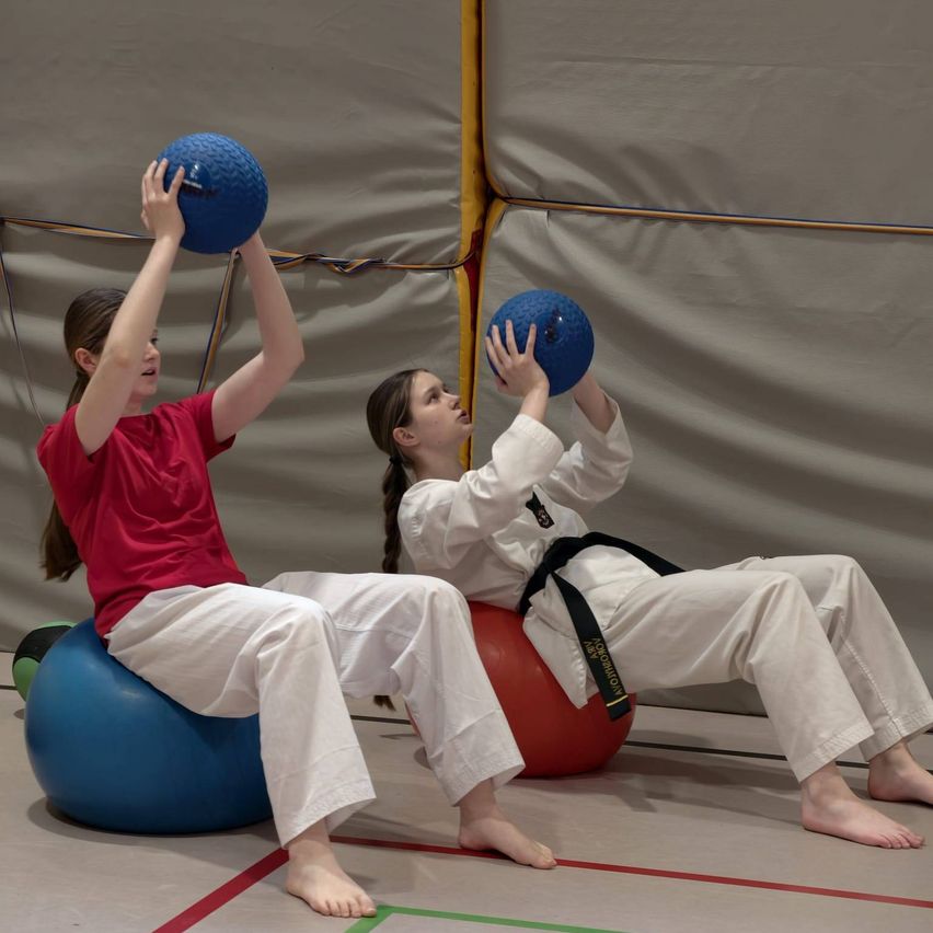 Zwei Frauen balancieren auf Übungsbällen. Eine hält einen blauen Ball, während die andere einen blauen Ball auf ihren Händen balanciert. Sie tragen alle weiße Hosen und weiße Oberteile.