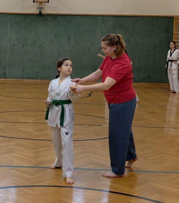 Eine Frau unterrichtet ein junges Mädchen in einer Karate-Uniform in einer Turnhalle. Eine andere junge Frau in einer Karate-Uniform steht im Hintergrund.