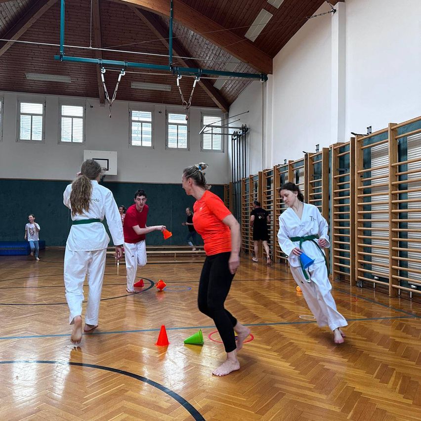Eine Gruppe von Menschen übt Kampfsport in einer Turnhalle. Sie sind barfuß, tragen weiße Uniformen und halten orangefarbene Kegel. Eine Frau springt und tritt einen Kegel.