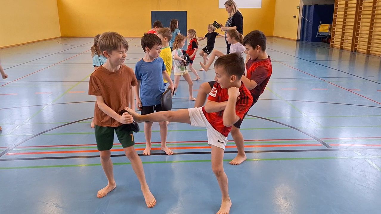 Eine Gruppe junger Jungen und Mädchen übt Kampfsport in einer Turnhalle. Einige sind barfuß, während andere Turnschuhe tragen. Eine Frau hält ein Buch und beobachtet sie.