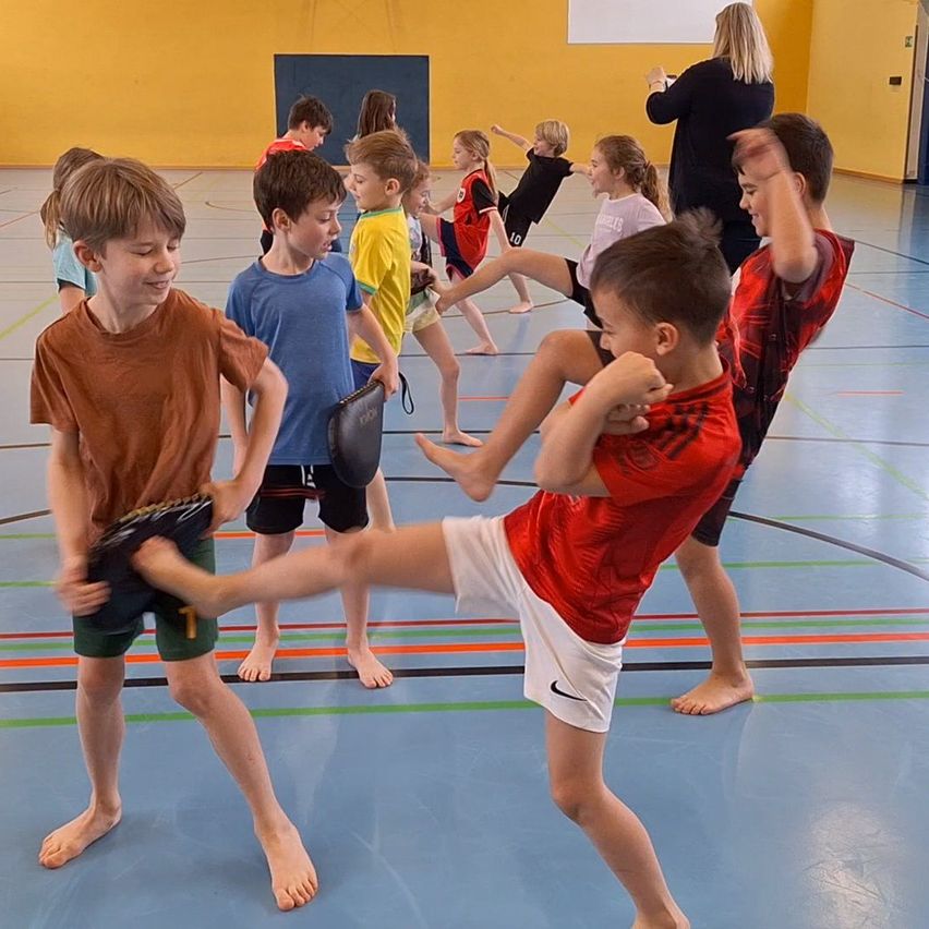Mehrere junge Kinder üben Kampfsport in einer Turnhalle. Einige sind barfuß, andere tragen Turnschuhe. Eine Frau beobachtet sie im Hintergrund.