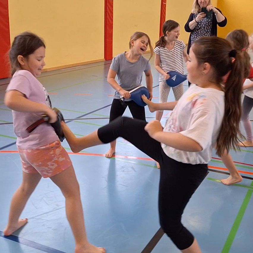 Mehrere Mädchen üben Kampfsport in einer Turnhalle, tragen Handschuhe und halten Pads. Ein Mädchen ist mitten in einem Tritt, während ein anderes ein Pad hält. Eine Frau im Hintergrund macht ein Foto.