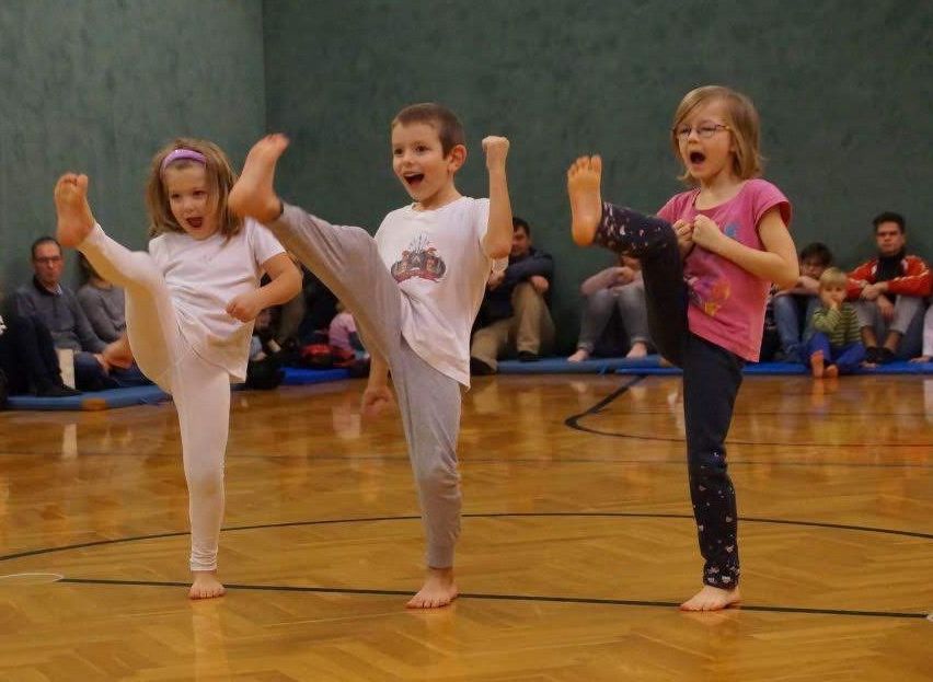 Drei Kinder führen Kampfsport-Kicks in einer Turnhalle aus. Der Junge in der Mitte trägt ein weißes T-Shirt, während die Mädchen rosa und weiße T-Shirts tragen. Sie sind barfuß und lächeln.
