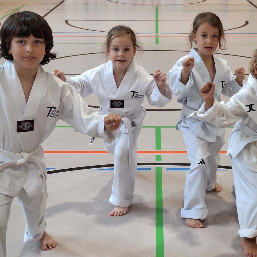 Vier Kinder in weißen Kampfsportuniformen stehen in einer Reihe und üben Karate-Posen auf einem Sporthallenboden mit mehrfarbigen Markierungen.