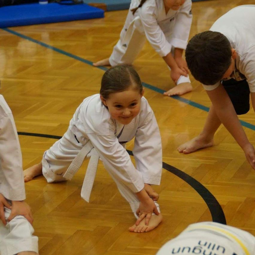 Eine Gruppe von Kindern in weißen Uniformen übt Kampfsport auf einem Holzboden. Ein Mädchen kniet und lächelt, während andere in verschiedenen Positionen sind.