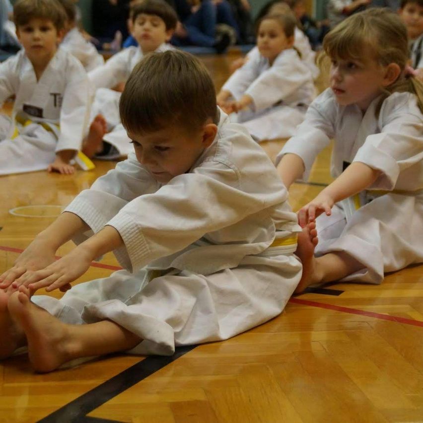 Eine Gruppe von Kindern in weißen Uniformen sitzt auf dem Boden und praktiziert möglicherweise Kampfsport. Der Junge vorne berührt seine Zehen.