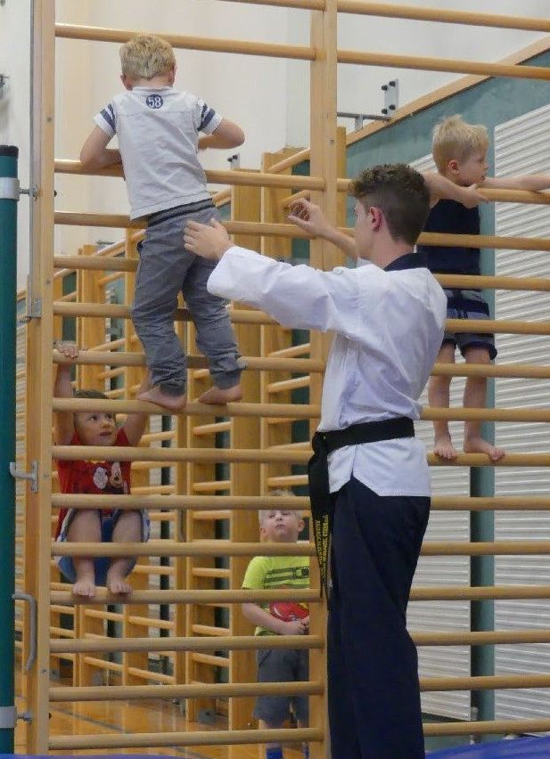 Ein Mann in einem weißen Hemd und einem schwarzen Gürtel hilft vier Kindern, eine Holzhindernisbahn zu erklimmen.