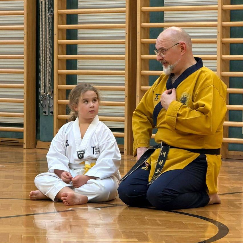 Ein älterer Mann in einem gelben Karate-Anzug kniet auf dem Boden und unterrichtet ein junges Mädchen in einem weißen Karate-Anzug. Sie sitzen beide auf dem Boden in einer Turnhalle.