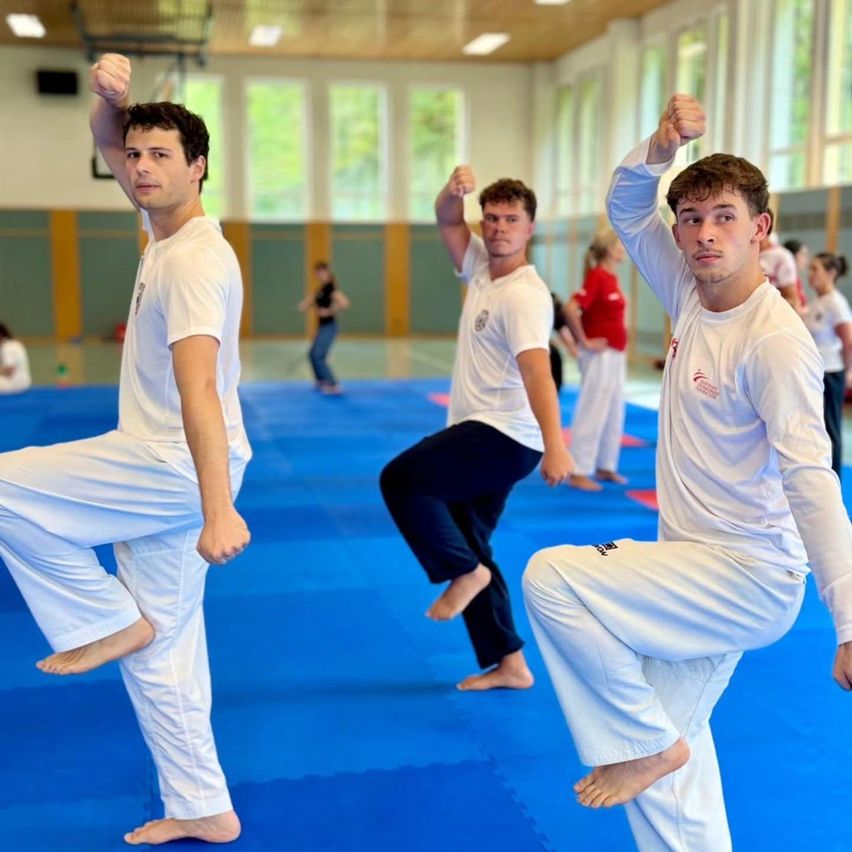 Drei Männer in weißen Uniformen und Hosen üben Kampfsport in einer Turnhalle. Der Mann auf der linken Seite befindet sich in einer Ausfallposition, während die anderen in einer Kampfhaltung sind. Im Hintergrund üben andere Menschen Kampfsport.
