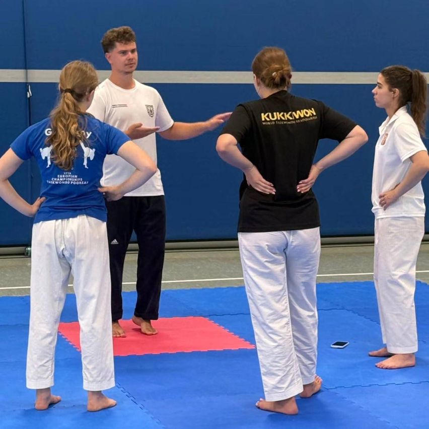 Vier Personen in Kampfsportkleidung stehen auf einem blauen Matten, ein Mann in einem weißen T-Shirt gestikuliert und spricht zu drei Frauen. Die Frauen haben ihre Hände auf dem Rücken, und ein Handy liegt auf dem Boden.
