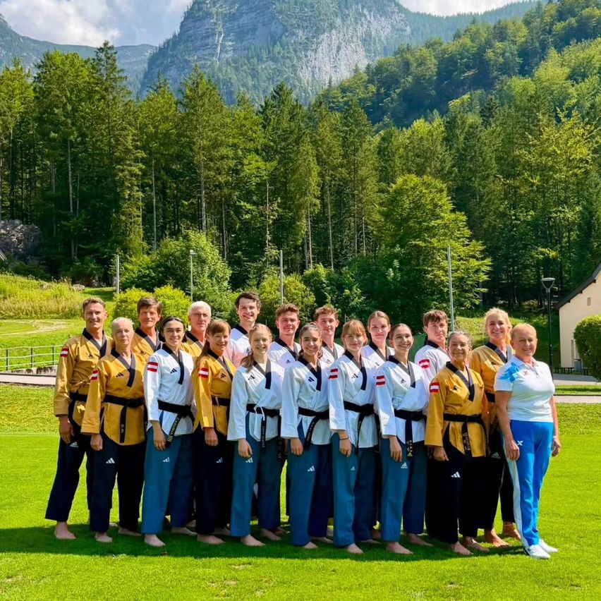 Eine Gruppe von Kampfsportlern in gelben und weißen Uniformen posiert für ein Foto auf einer Rasenfläche mit einer Kulisse aus Bergen und Bäumen.