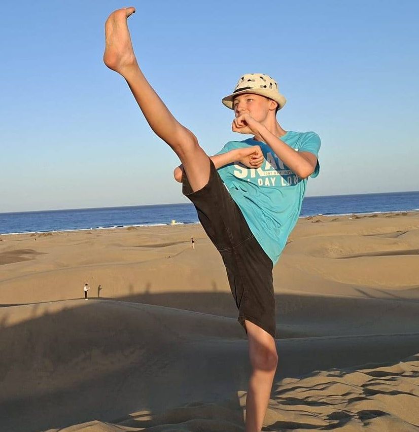 Ein junger Junge führt einen hohen Kick auf einem sandigen Strand aus, mit einem klaren blauen Himmel im Hintergrund.