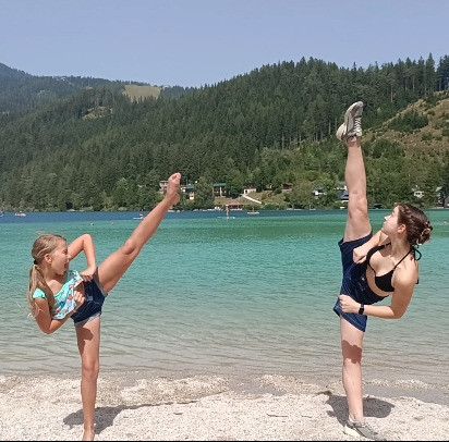 Zwei Frauen üben Kampfsport-Kicks an einem Sandstrand mit einem See und Bergen im Hintergrund.