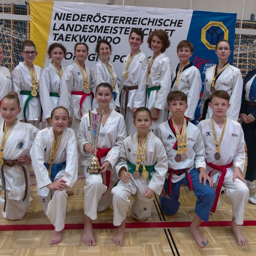 Eine Gruppe junger Taekwondo-Athleten, die weiße Uniformen und Medaillen tragen, posiert für ein Foto vor einem Banner. Sie lächeln und scheinen einen Sieg zu feiern.