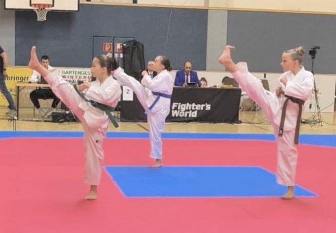 Zwei junge Mädchen in pinkfarbenen Kampfsportuniformen führen hohe Tritte in einer Turnhalle aus. Im Hintergrund steht ein Tisch mit einem Schild und einem Mann in einem blauen Anzug.