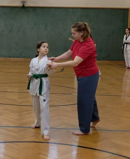 Ein junges Mädchen in einem weißen Karate-Anzug mit einem grünen Gürtel wird von einer Frau in einem roten Shirt in einer Turnhalle geführt.