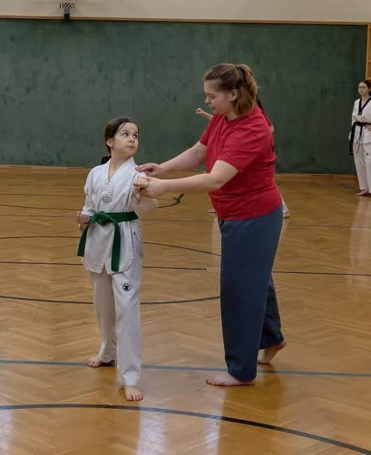 Ein junges Mädchen in einem weißen Karate-Anzug mit einem grünen Gürtel wird von einer Frau in einem roten Shirt in einer Turnhalle geführt.