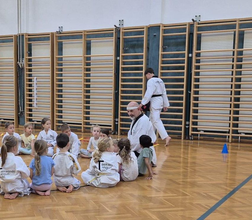Eine Gruppe junger Kinder in weißen Uniformen sitzt in einem Fitnessstudio auf dem Boden und hört einem Mann in weiß zu. Hinter ihnen geht ein Mann.