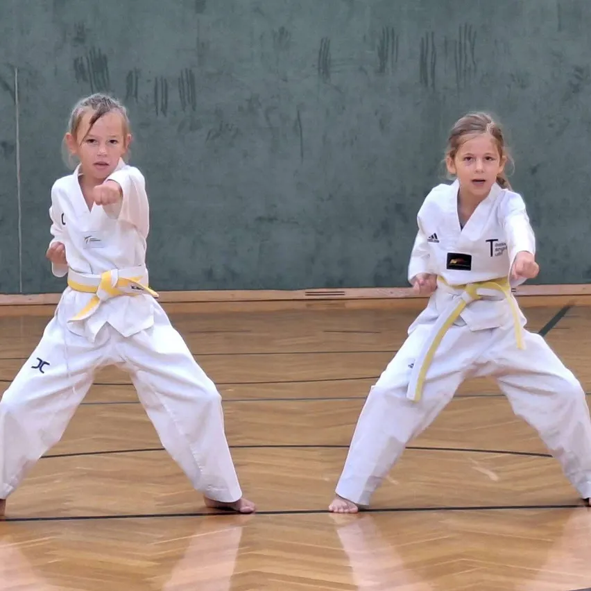 Zwei junge Mädchen in weißen Karate-Uniformen mit gelben Gürteln führen Kampfkunst-Posen auf einem Holzboden aus.