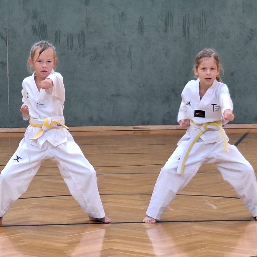 Zwei junge Mädchen in weißen Karate-Uniformen mit gelben Gürteln führen Kampfkunst-Posen auf einem Holzboden aus.