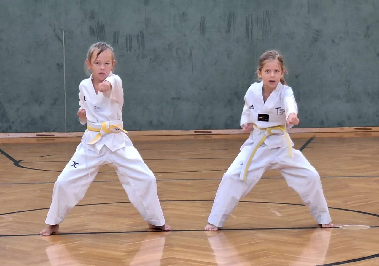 Zwei junge Mädchen in weißen Karate-Uniformen mit gelben Gürteln führen Kampfkunst-Posen auf einem Holzboden aus.