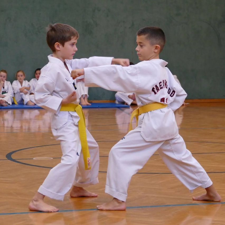 Zwei junge Jungen in Karate-Uniformen üben Kampfsport auf einem Platz. Ein Junge hat einen gelben Gürtel, und sie sind in einem Sparring-Match engagiert.