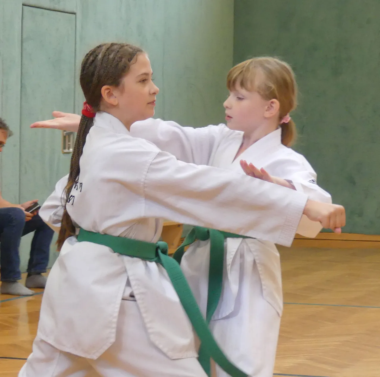 Zwei junge Mädchen in weißen Karate-Uniformen mit grünen Gürteln üben Kampfkunst in einer Turnhalle. Ein Mädchen hält den Arm des anderen.