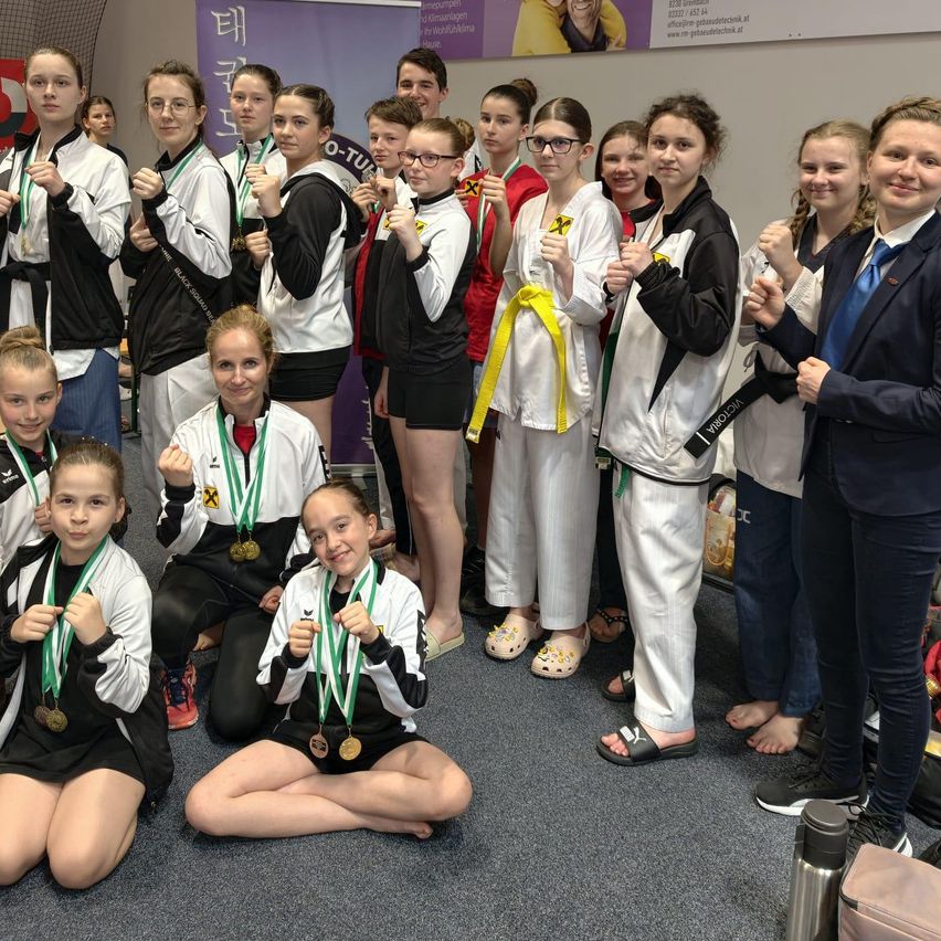 Eine Gruppe junger Menschen in Kampfsportuniformen posiert für ein Foto, mit Medaillen um den Hals. Sie lächeln und stehen eng beieinander.