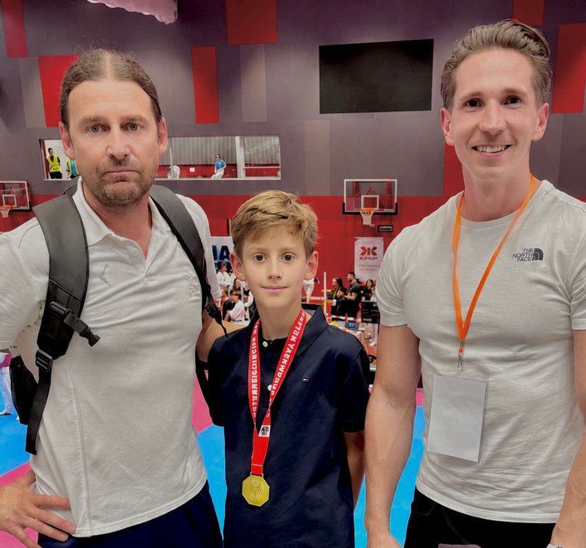 Drei Personen stehen gemeinsam in einem Fitnessstudio. Ein Junge mit einer Medaille um den Hals steht zwischen zwei Männern. Die Männer tragen weiße Hemden mit orangefarbenen Lanyards.
